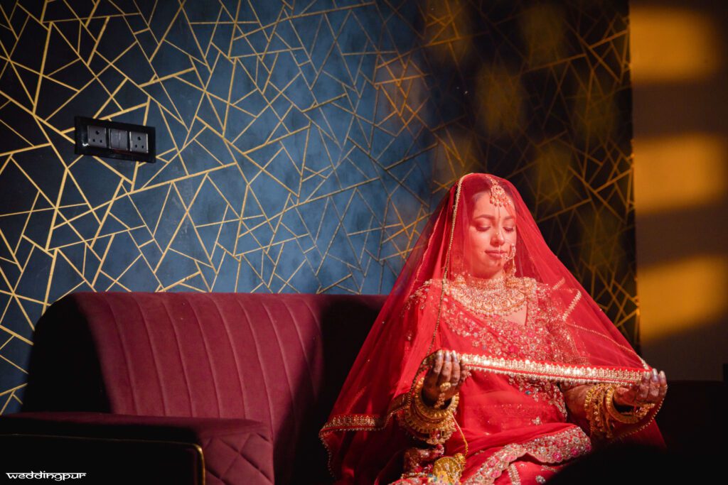 Bride sitting on sofa holding veil in red lehenga with warm lighting
